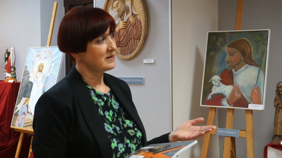 Danuta Wachowska, kobieta prezentująca prace na wystawie w tle obrazy z Chrystusem