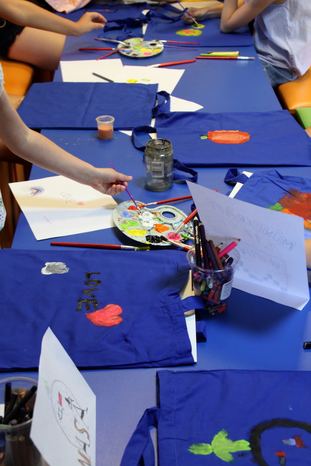 Na zdjęciu widoczne są granatowe torby leżące na stolikach. Dzieci malują farbami torebki dla najbliższych