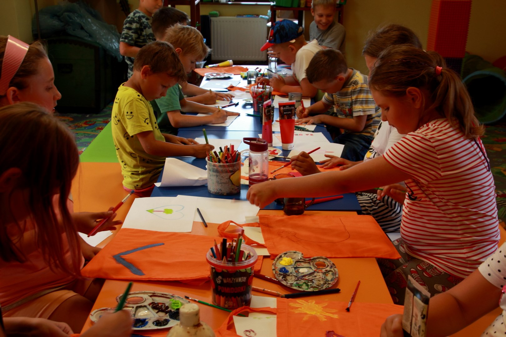 Na zdjęciu grupa dzieci , chłopcy i dziewczynki siedzący przy stole. Przed nimi leżą kredki, palety w z farbami oraz tuby farb. Dzieci malują wzory na pomarańczowych torbach