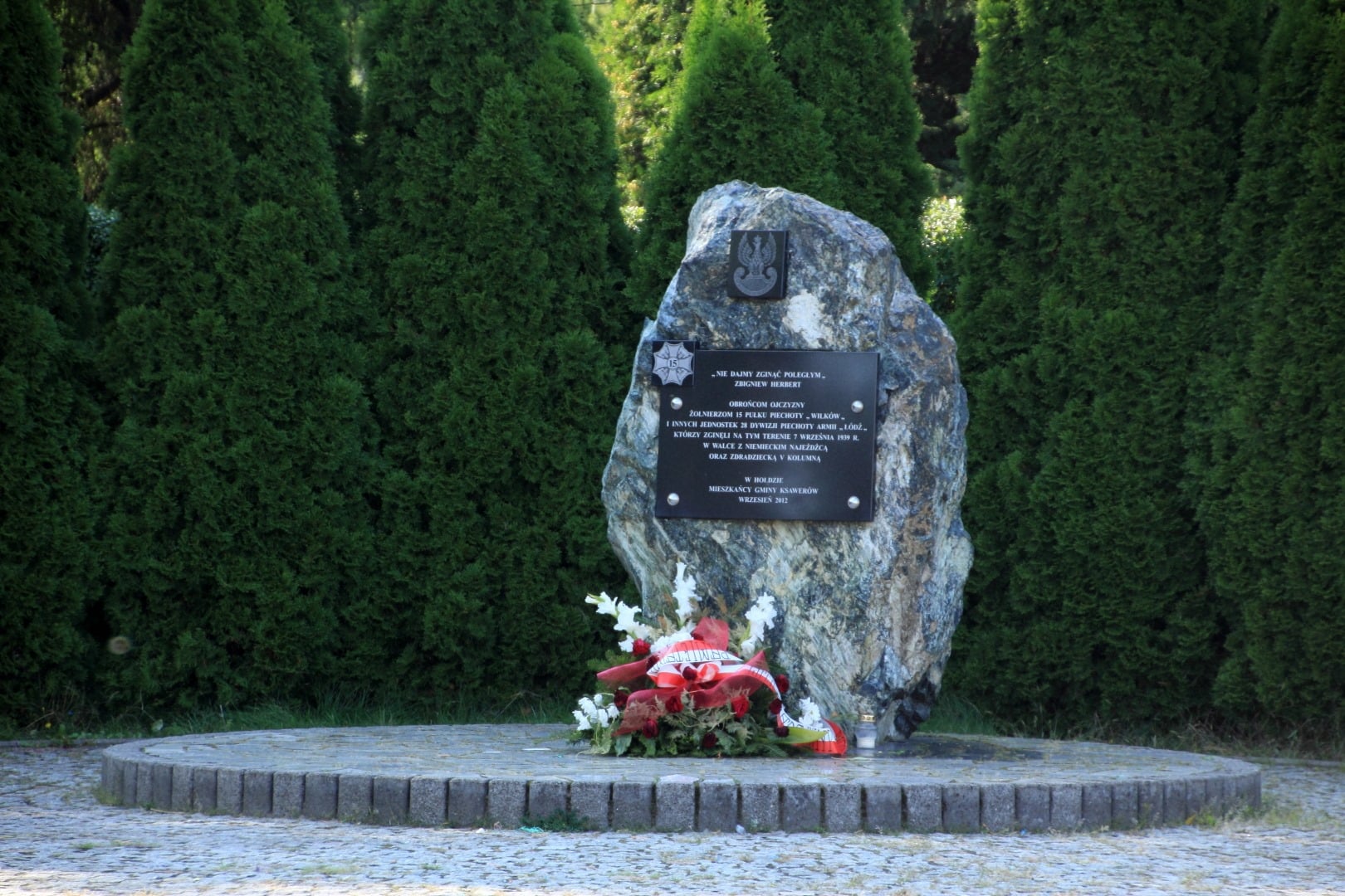 Zdjęcie przedstawia obelisk (kamień) przed którym znajduje się wieniec biało czerwonych kwiatów