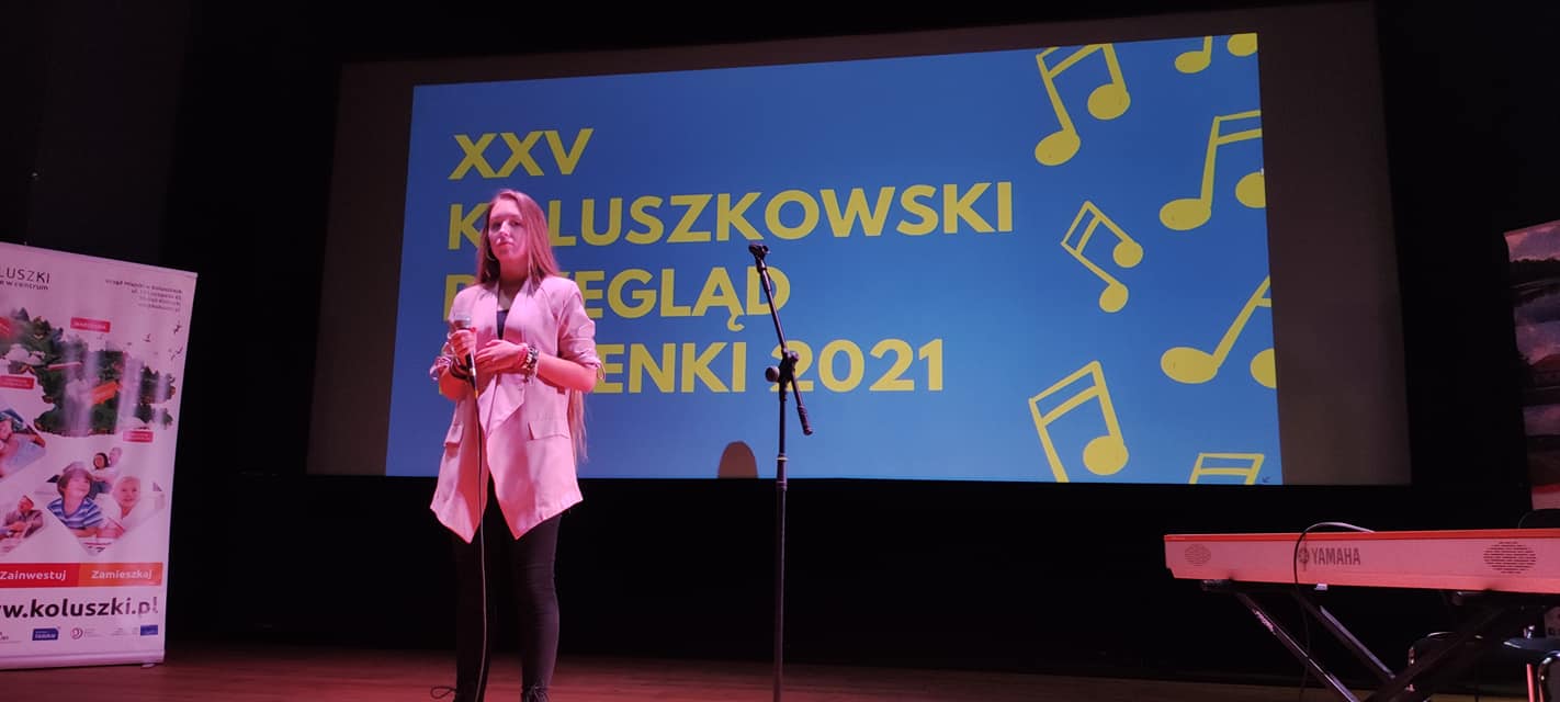 Młoda kobieta na scenie z mikrofonem. W tle napis" XXV Koluszkowski Przegląd Piosenki 2021