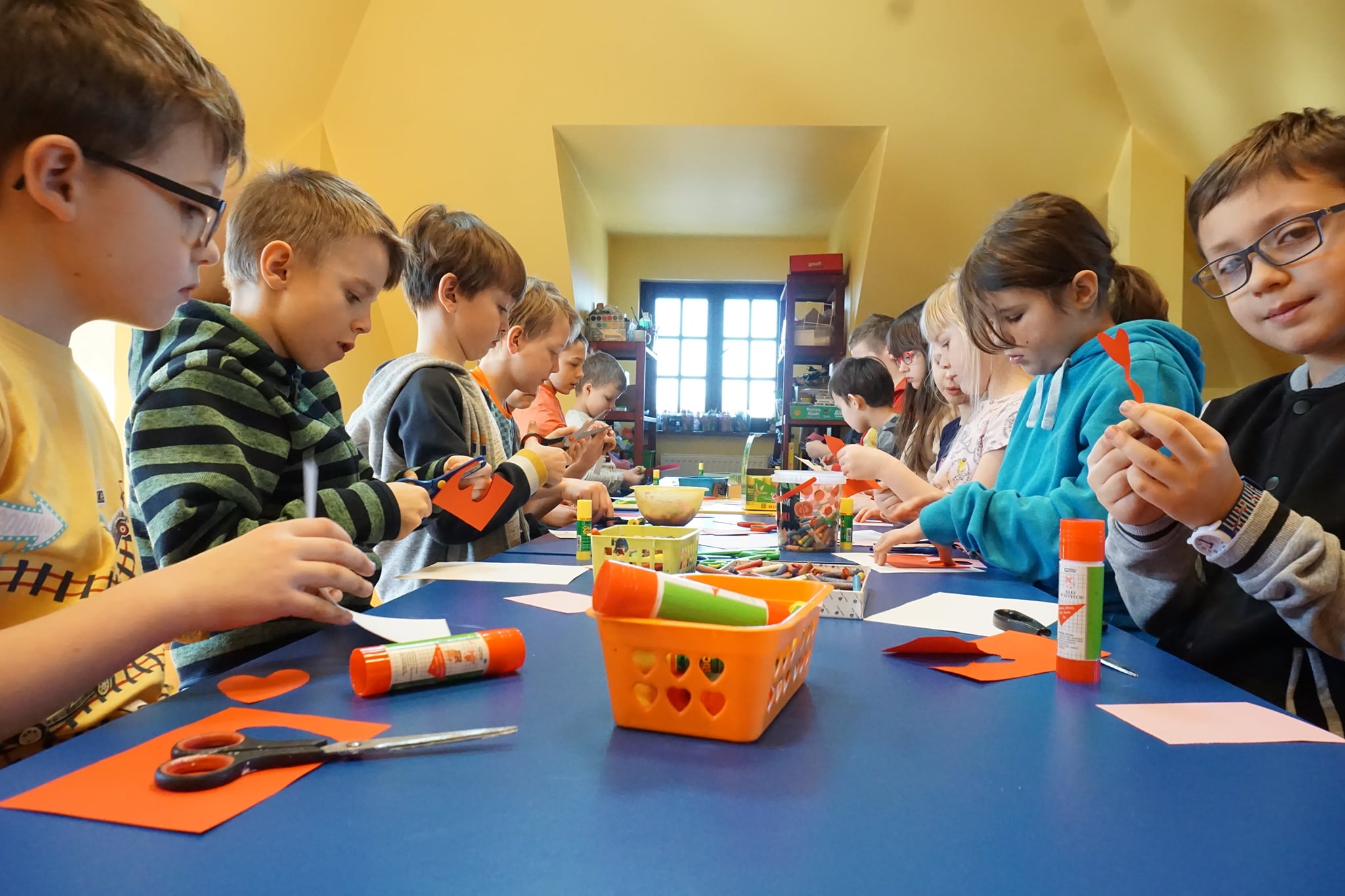 Grupa dzieci, chłopcy i dziewczynki siedzący przy stole, wycinający serduszka na kartki walentynkowe