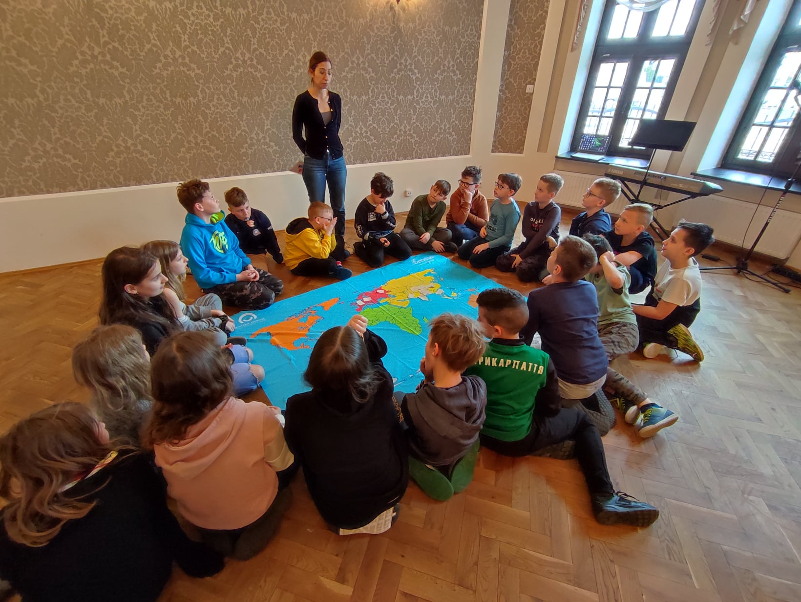 Uczestnicy warsztaów wraz z prowadzącą. Dzieci siedzą w kole wokół mapy świata. Kobieta prowadząca zajęcia stoi poza kołem