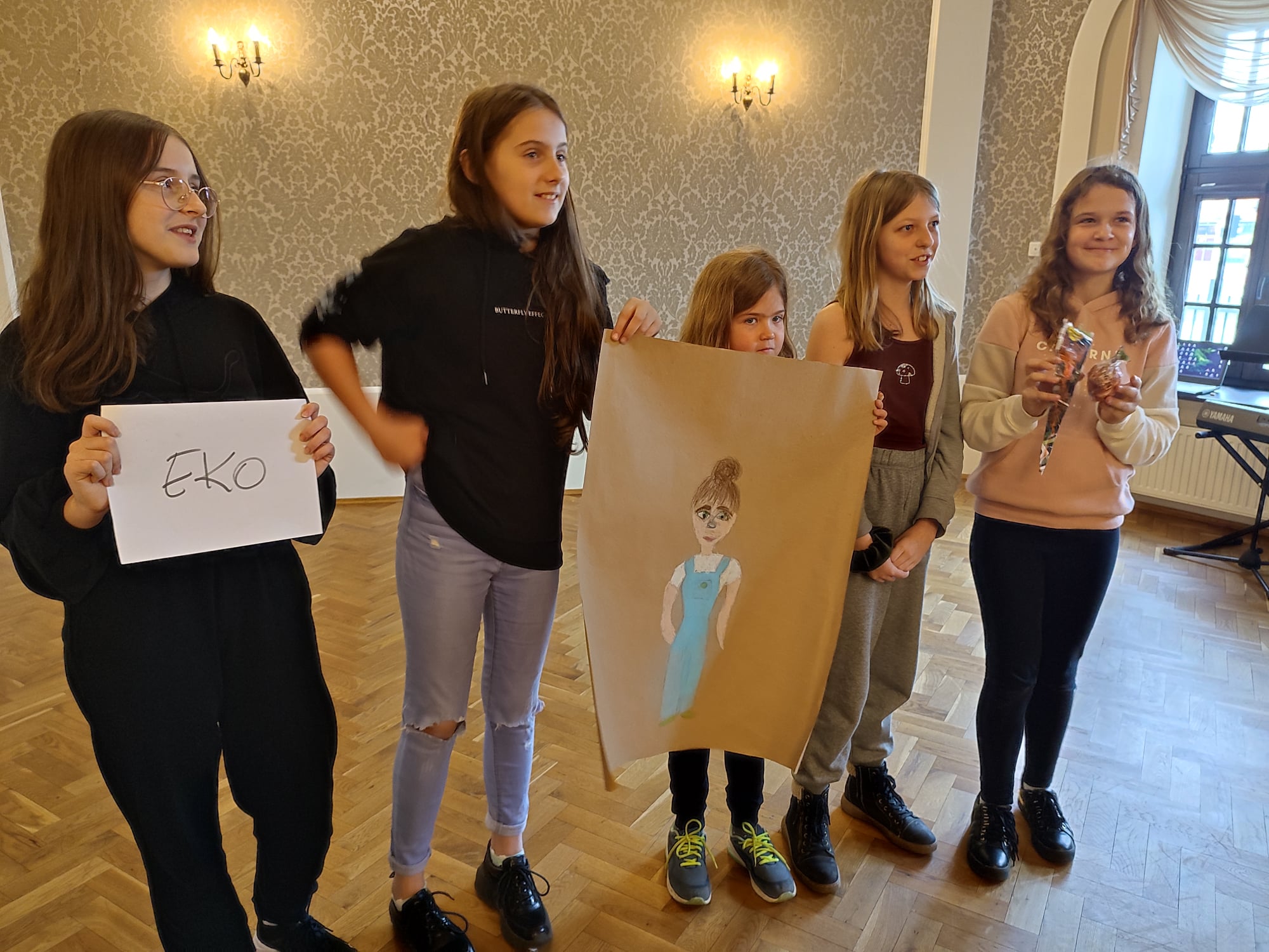 Pięć dziewczynek prezentuje swój plakat, z postacią w niebieskiej sukience. Jedna osoba trzyma kartkę z napisem eko