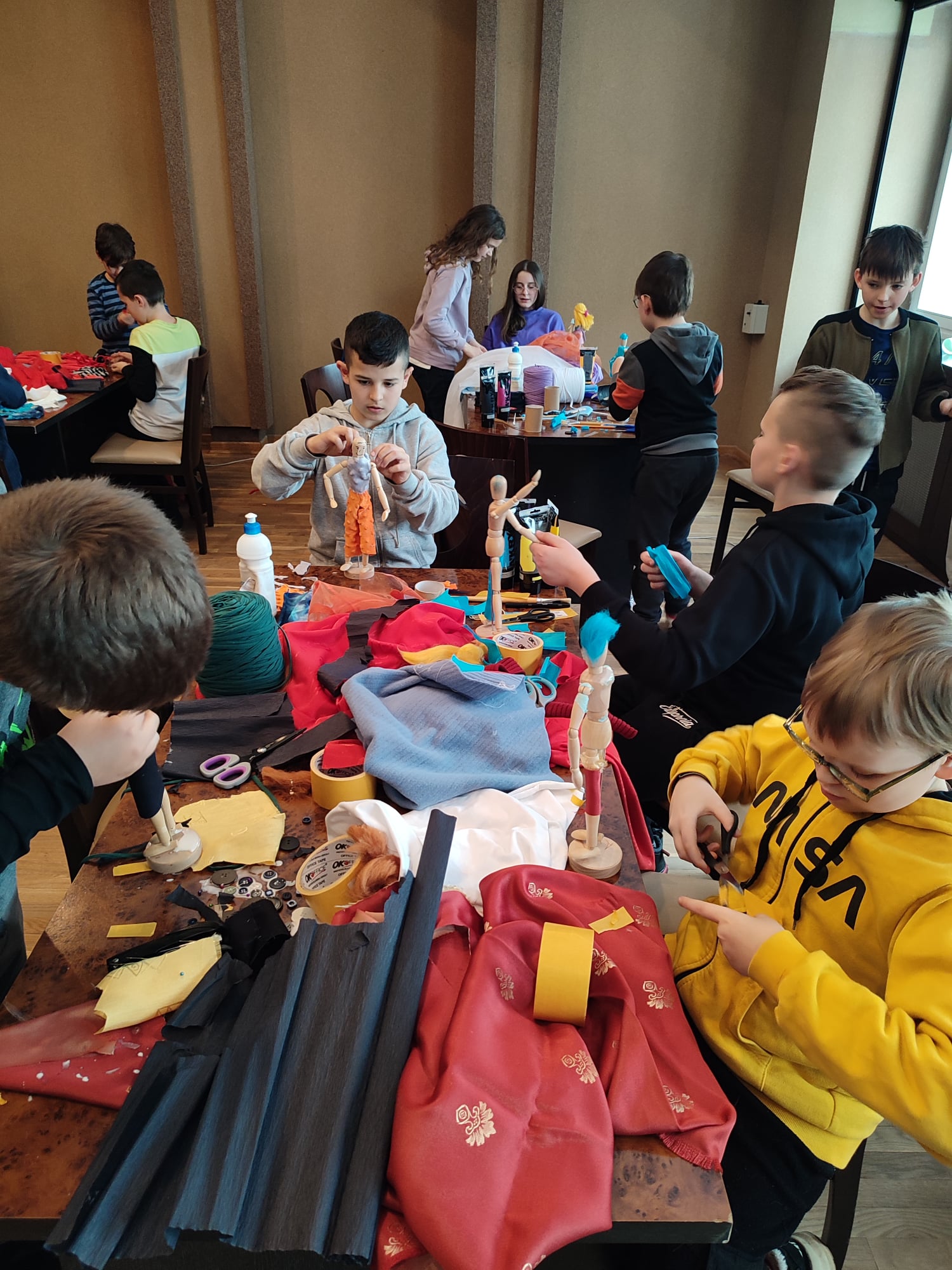 Grupa dzieci, chłopcy i dziewczynki, siedzący przy stolikach, przed nimi manekiny, materiały kolorowe