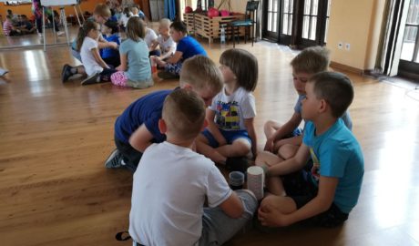 Dwie grupy dzieci, chłopcy i dziewczynk siedzą na podłodze w sali baletowej. w tle lustro z odbcie, dzieci