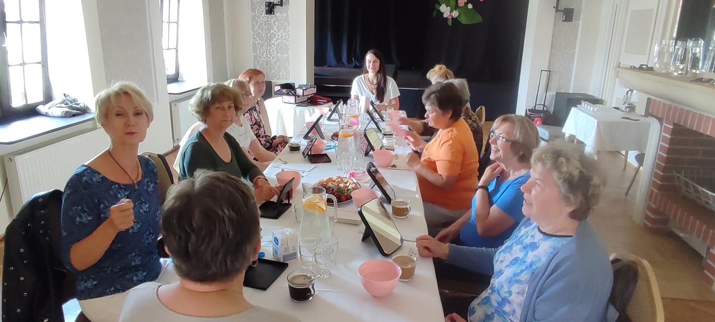 Dziesięć kobiet siedzących przy stole. Przed nm lusterka. Na stole ciastka, dzbanki z wodą. 