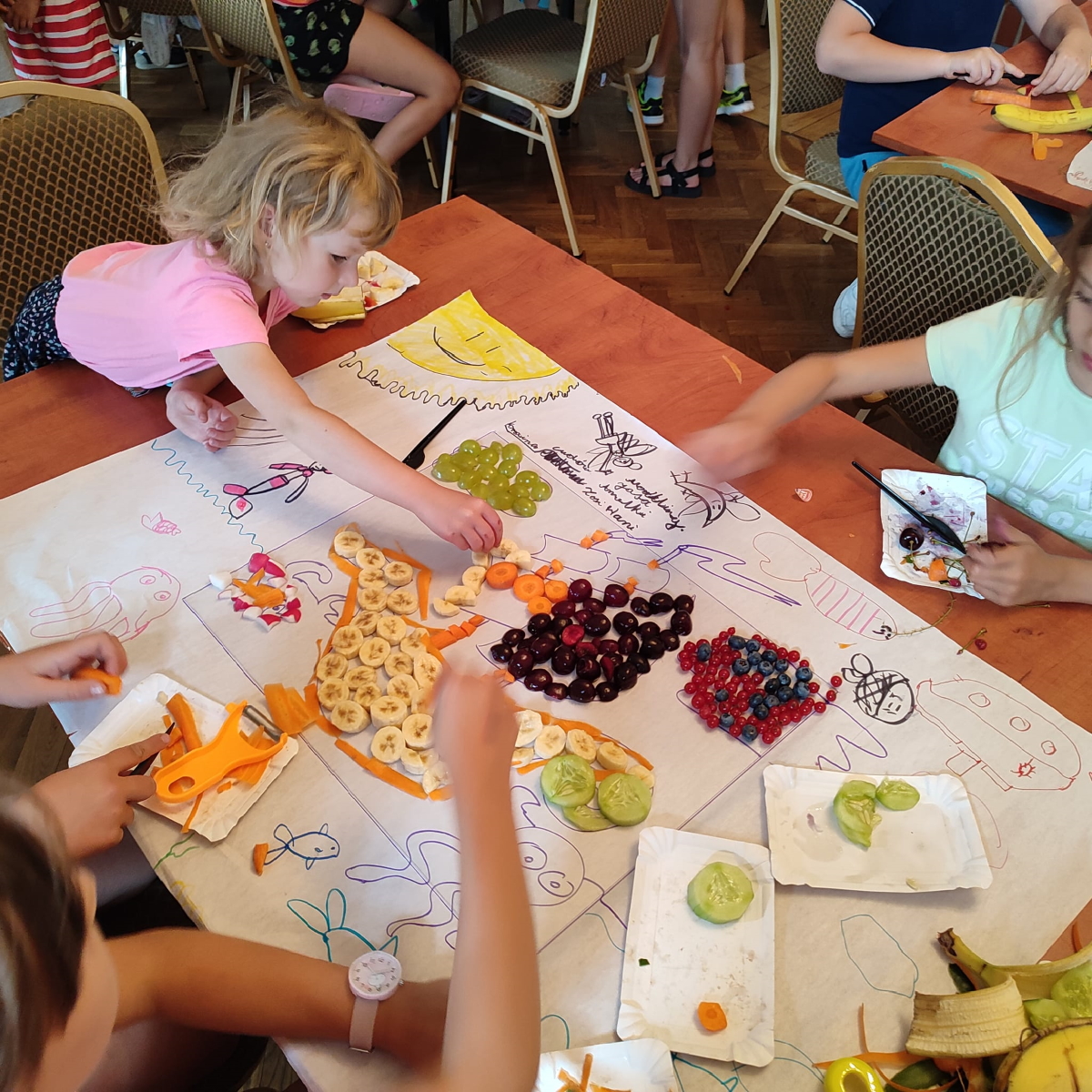 dziewczynki siedzące przy stolikach układaja obrazek z owoców