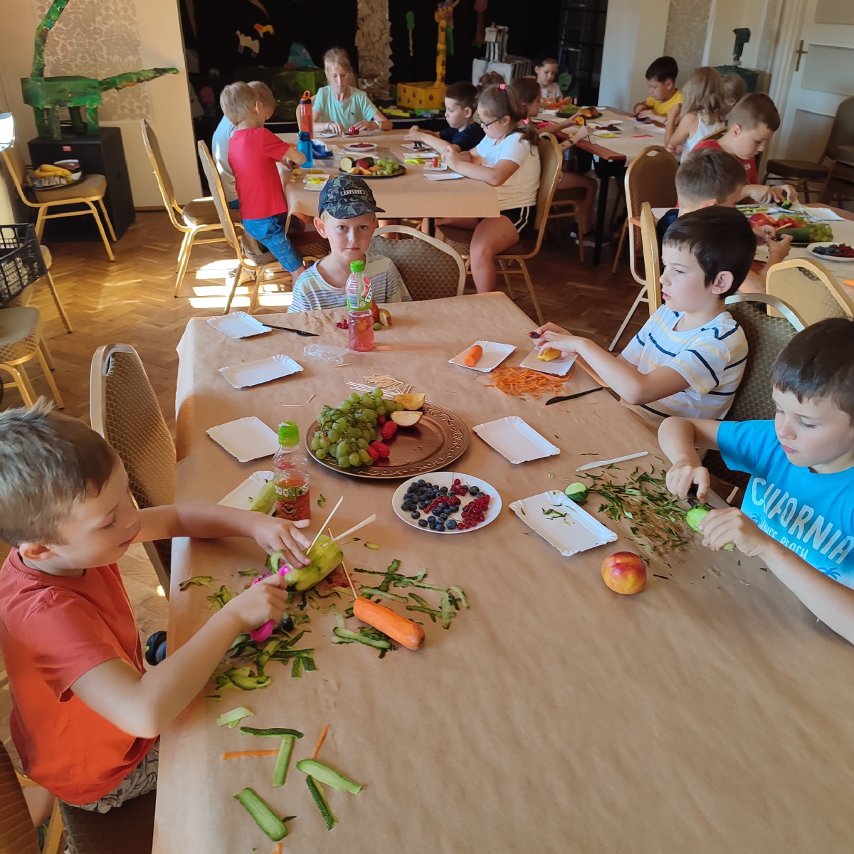 dzieci siedzące przy stolikach, przed nimi owoce i warzywa. Dzieci obierają warzywa: ogórki i marchewkę