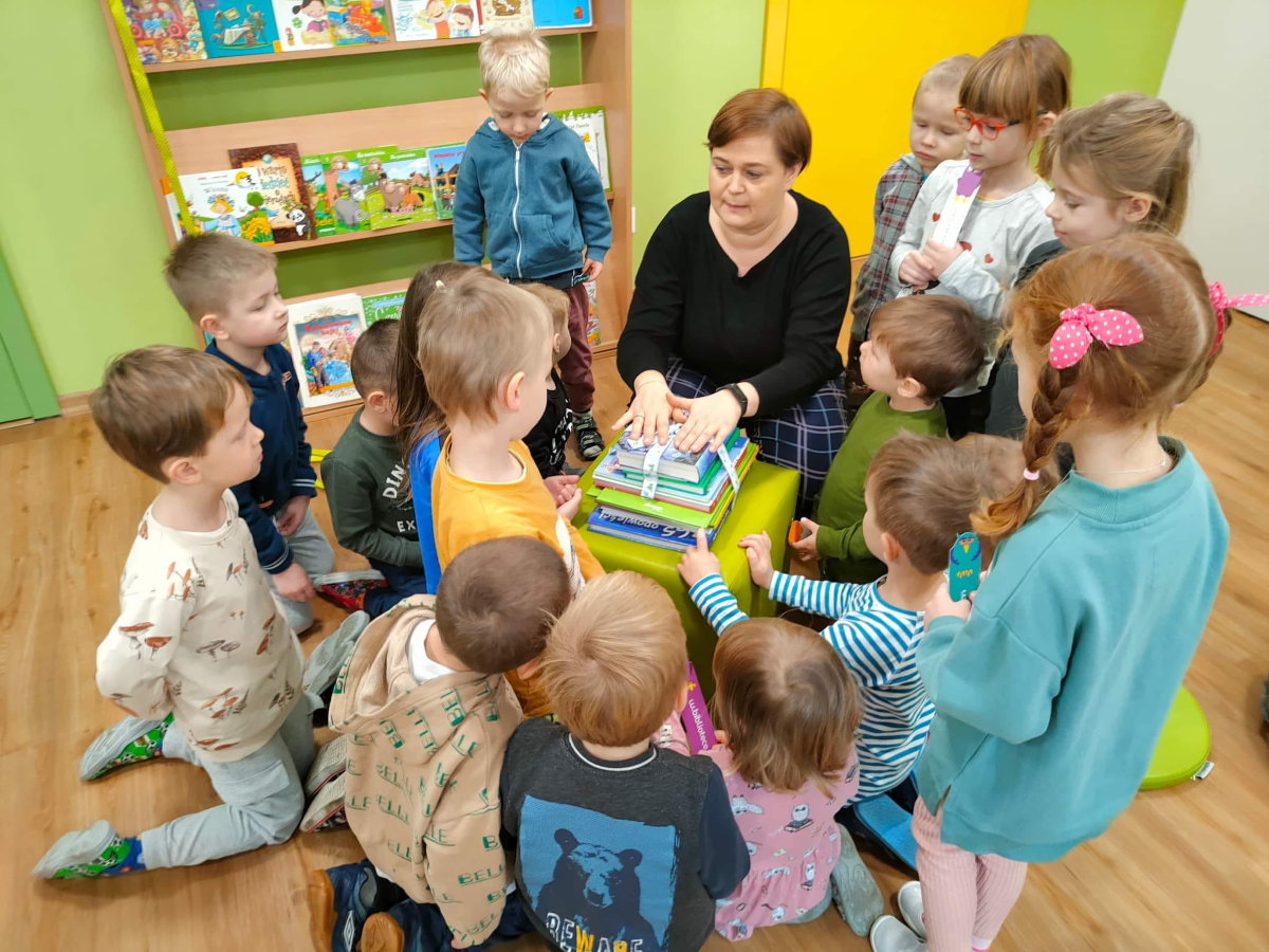 Pani Bibliotekarka siedzi przed grupą dzieci trzymając dłonie na książkach