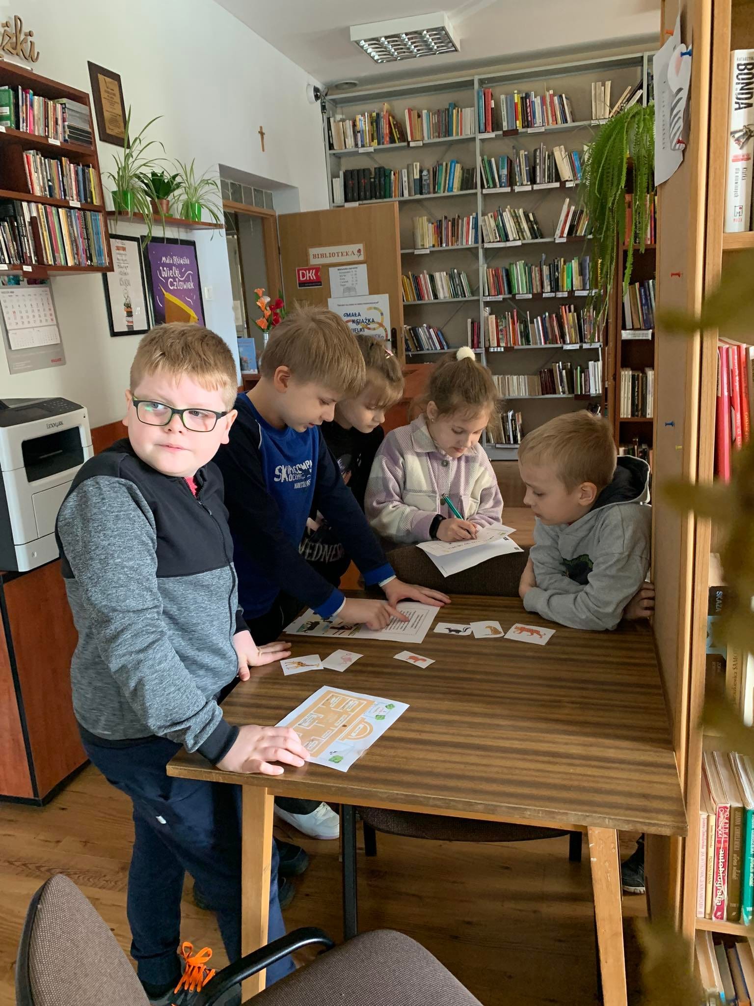 Piątka dzieci w bibliotece. Dzieci stoją przy stoliku i rozwiązują zadania