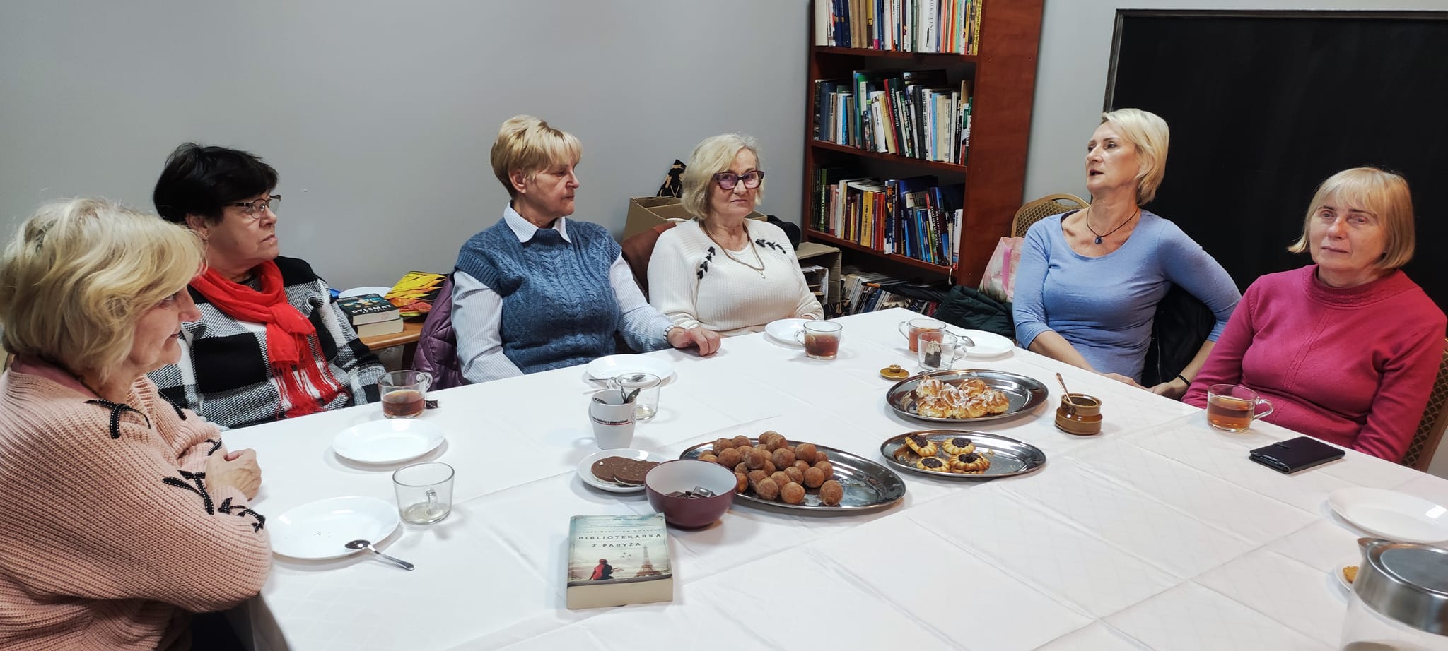 grupa kobiet siedzi przy stole. na stole ciasteczka, kubki z kawą oraz książka